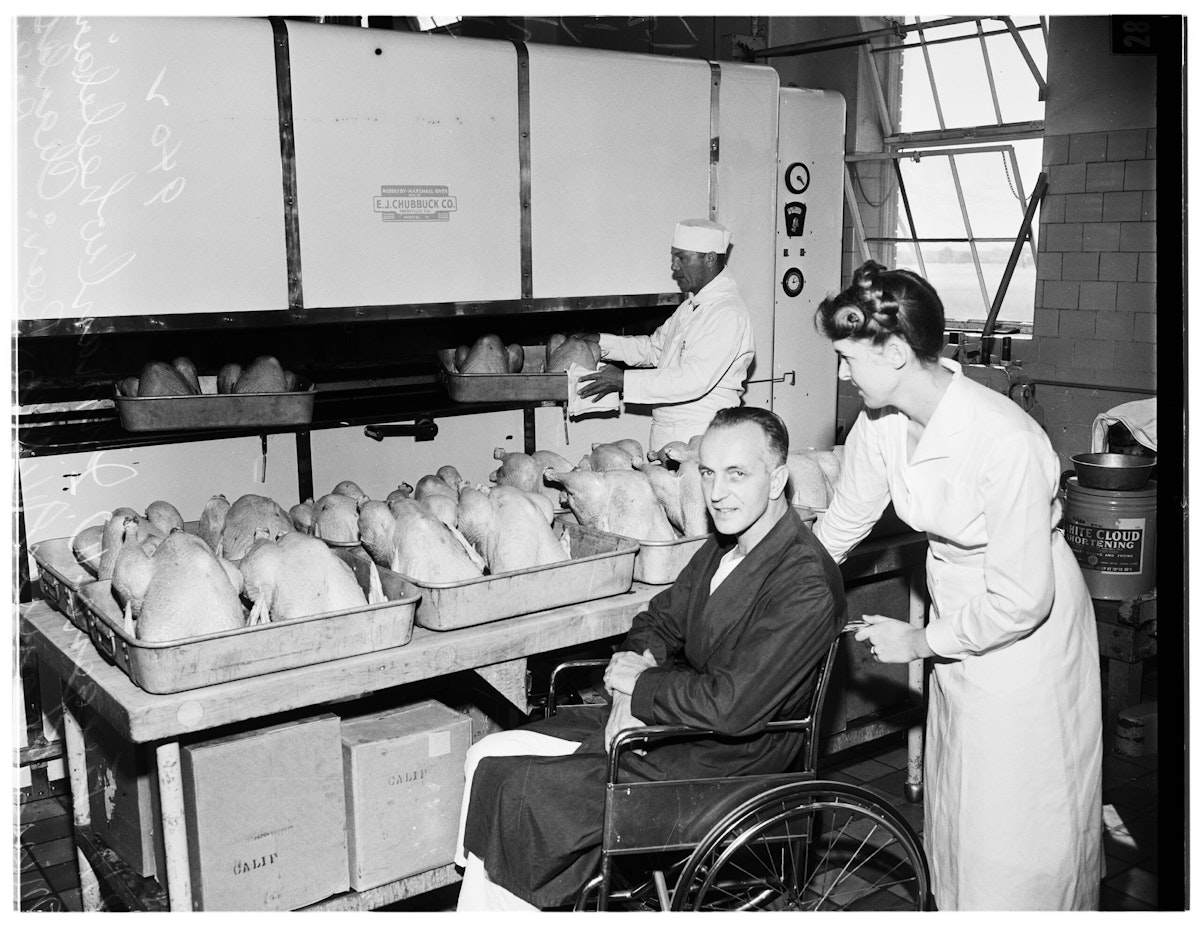 A nurse wheels a smiling man past several pans of turkeys being prepared for a Thanksgiving meal at the West Los Angeles Veterans Administration.