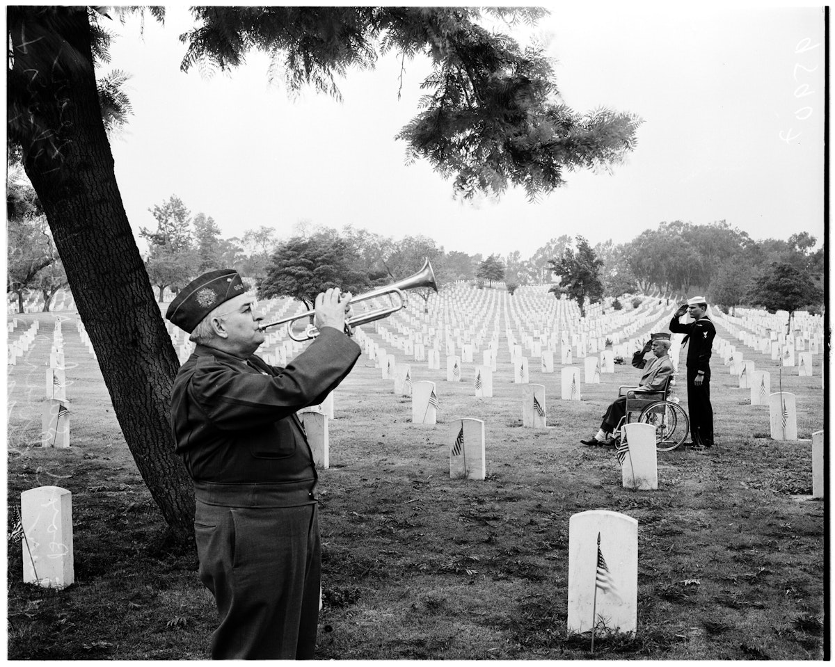 A veteran plays the trumpet in the Sawtelle Veterans Administration Center cemetery, as two other veterans salute in the background.