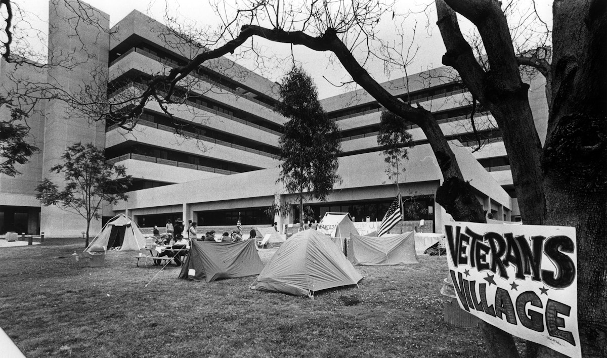 A sign that reads "Veterans Village" is posted on a tree next to a collection of tents occupied by veterans.