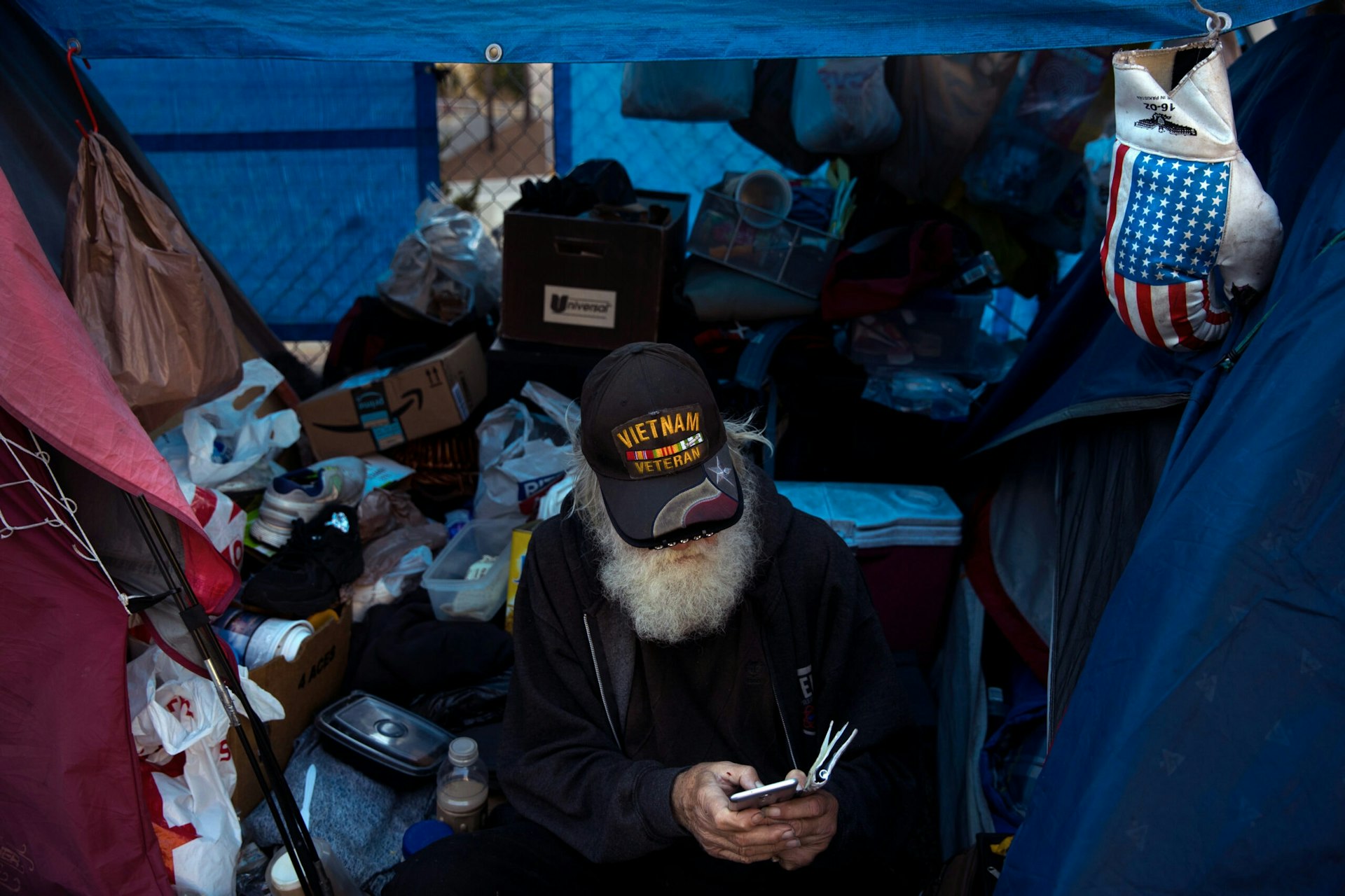 An elderly man, wearing a Vietnam veteran hat, looks at his cell phone in a homeless encampment.