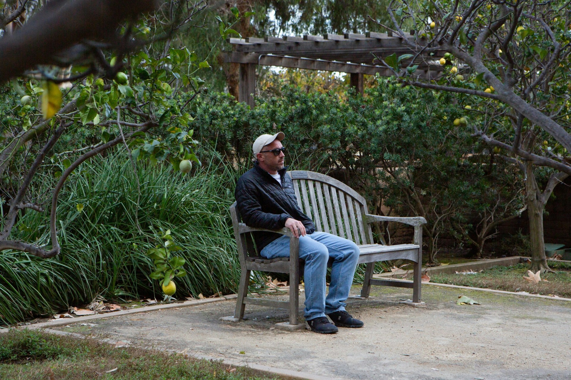 Rob Reynolds, wearing a baseball hat and sunglasses, sits on a wooden bench in the West LA VA rose garden.