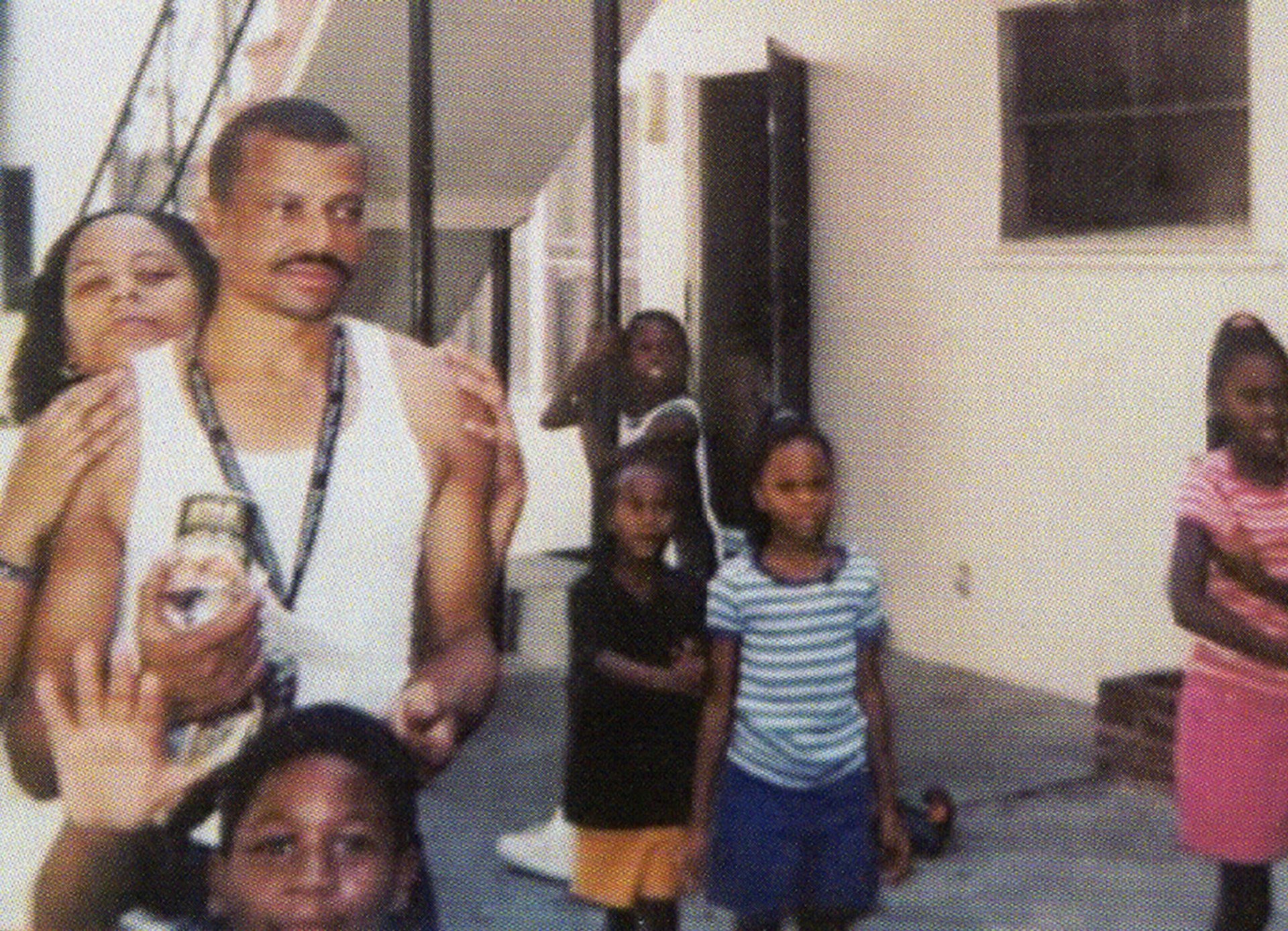 Andre Butler, as a young man, stands near a staircase, as a woman places her hands on his shoulders, and several children play nearby.