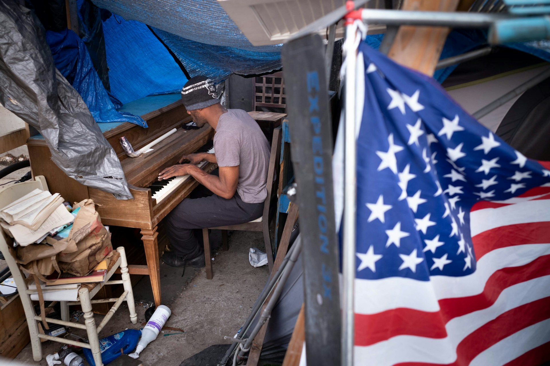 An unhoused man plays an upright piano in a makeshift tent, surrounded by his belongings and with an American flag in the foreground.