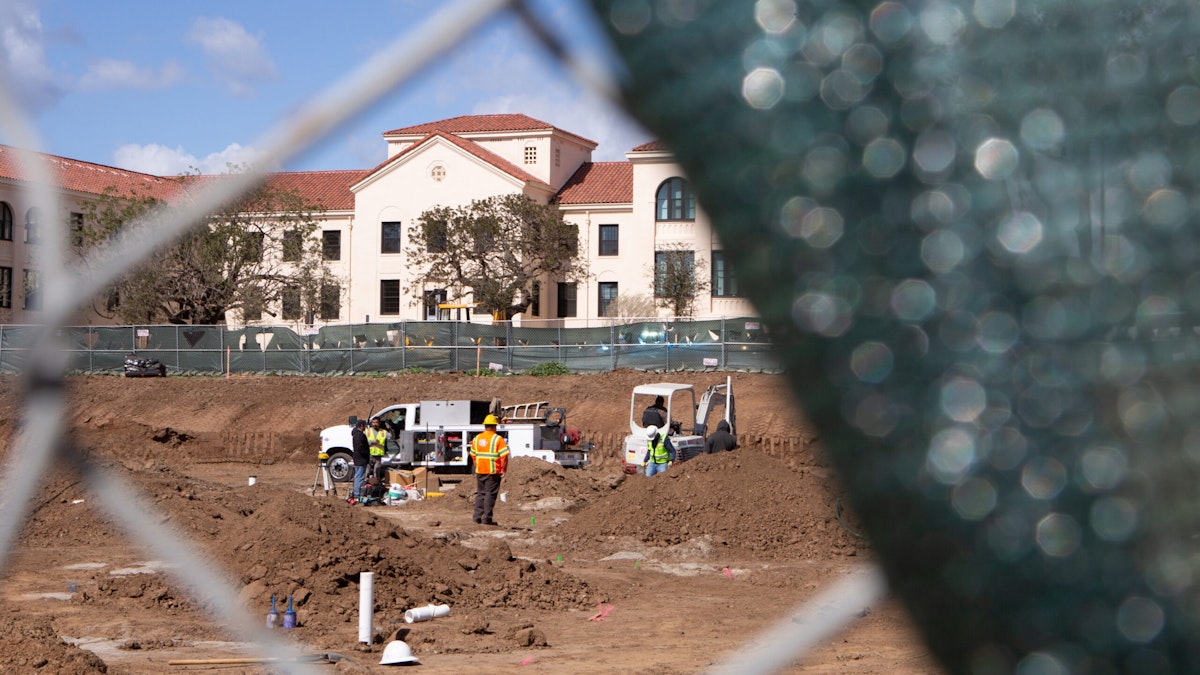 Construction workers are seen through a chain-link fence, with residential VA housing visible in the background.